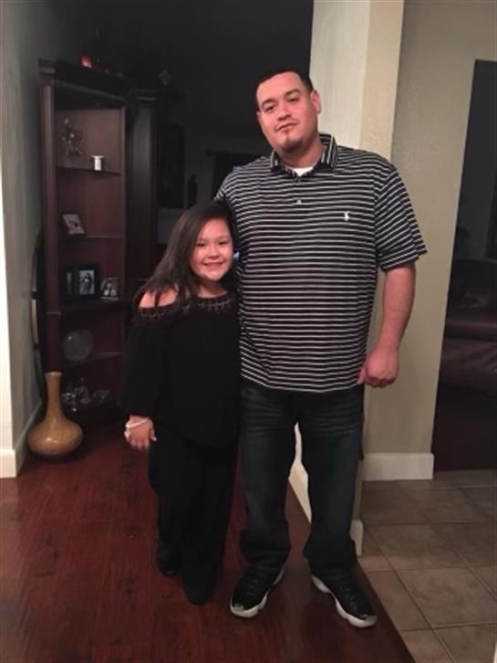 A father stands proudly with his daughter, both smiling in a warm living room setting.