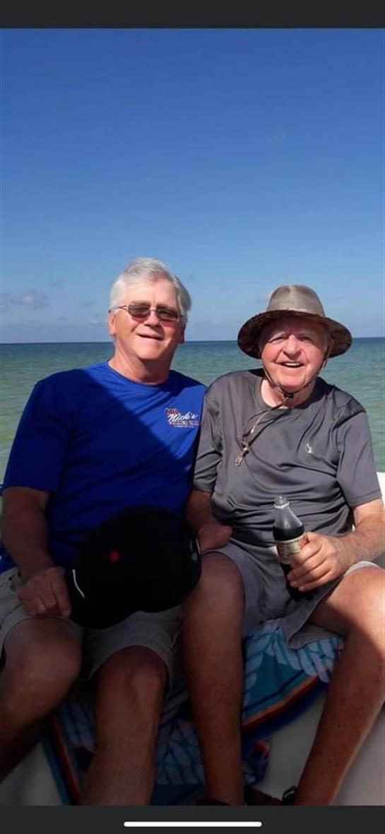 Two men sit together on a boat, smiling joyfully against a backdrop of calm water and blue sky.