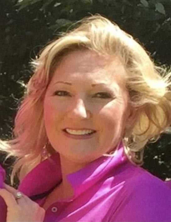 A woman with wavy blonde hair smiles brightly while standing outside on a sunny day.