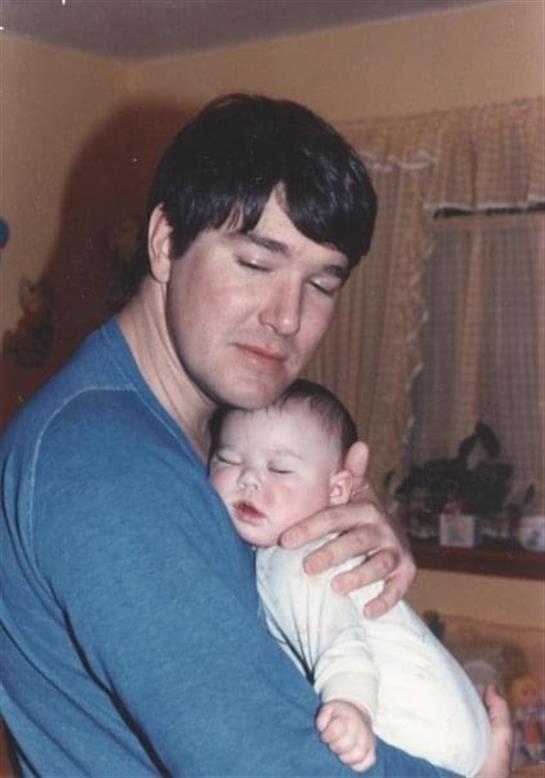 A father holds his sleeping baby close, expressing warmth in a vintage home environment.