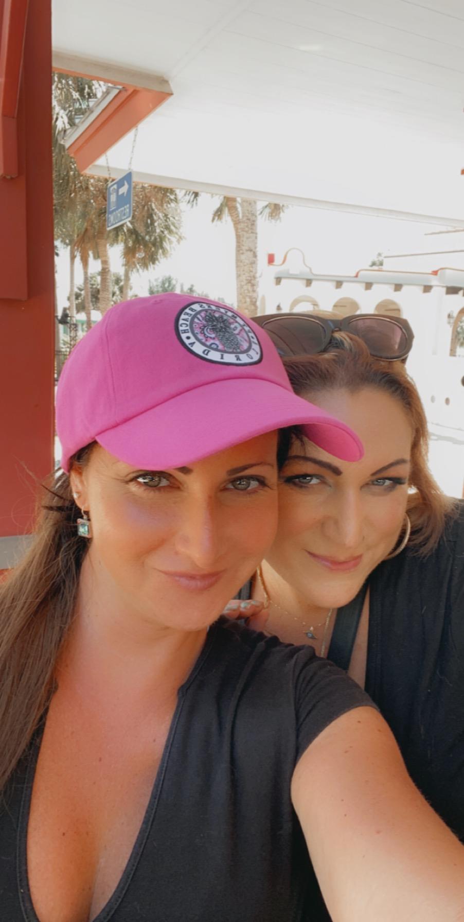 Two women smile brightly while taking a selfie in a lively outdoor setting filled with palm trees.