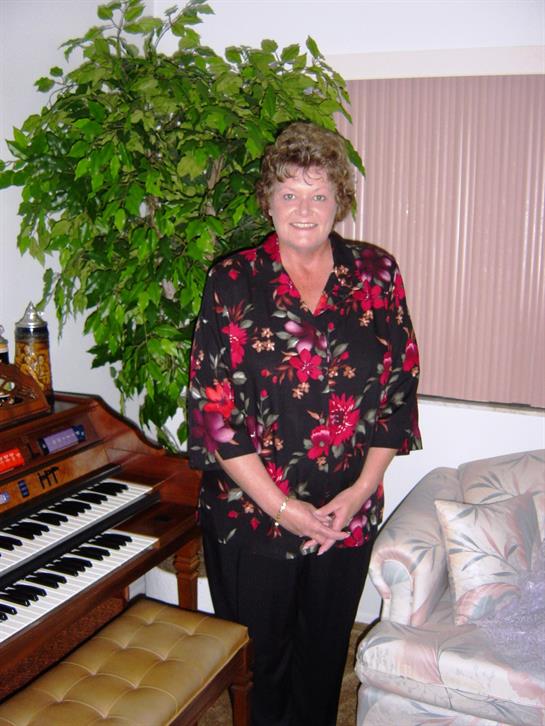 A woman poses next to a vintage organ surrounded by a vibrant indoor plant in a warm living space.