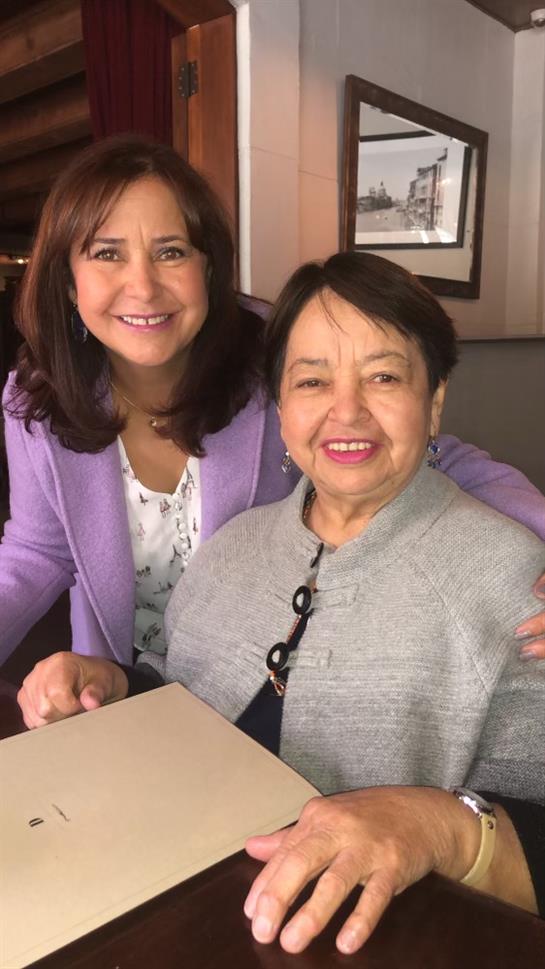 Two women smile warmly while sitting at a restaurant table, enjoying each other’s company.