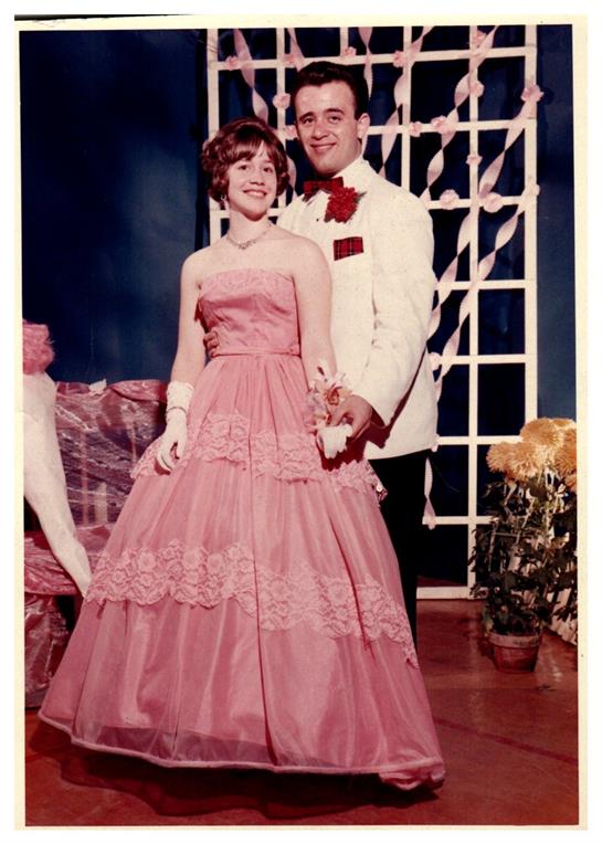Elegant couple poses in formal attire at a dance surrounded by decorations and flowers.