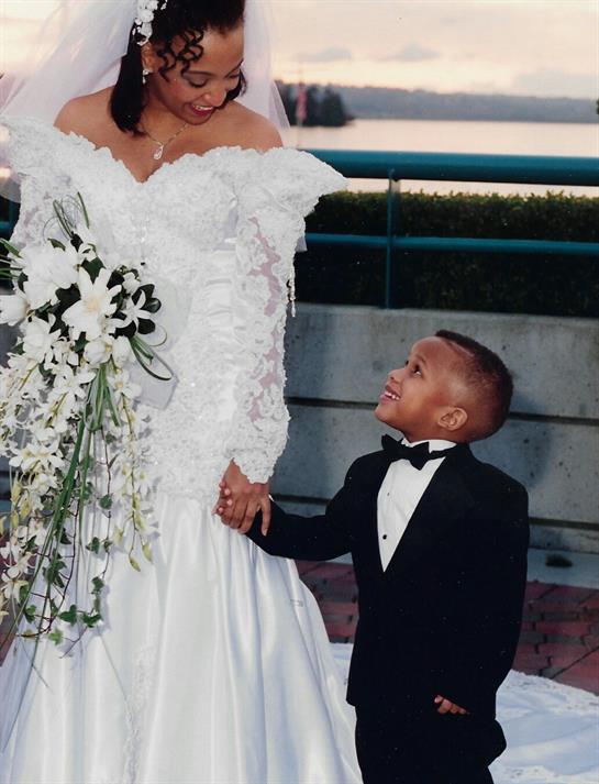 Couple exchanges smiles at wedding ceremony while child holds bride's hand, outdoors.