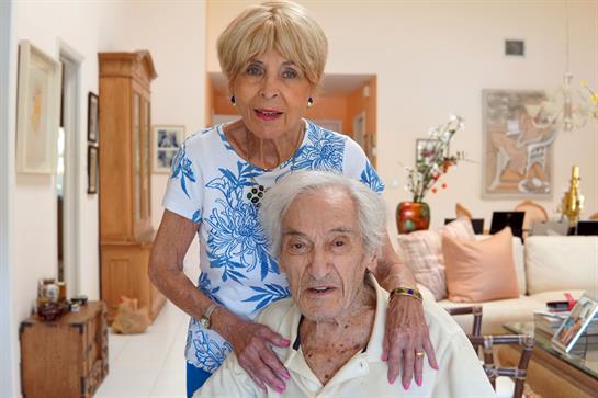 An elderly couple enjoys each other's company in their bright living room, smiling warmly together.