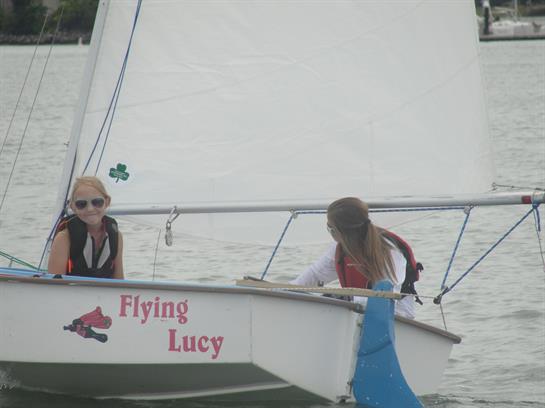 Two girls on a sailboat, smiling and participating in a sailing activity on a lake.