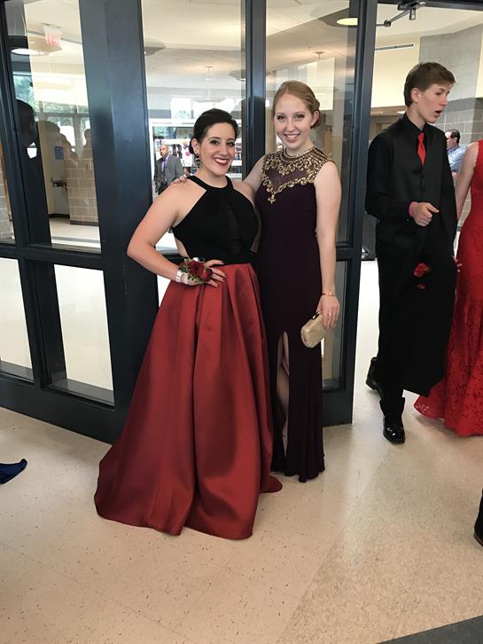 Two friends pose joyfully in stunning formal dresses at a lively social gathering.