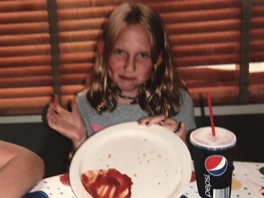 Child smiles while holding an empty plate with remnants of ketchup, seated at a table.