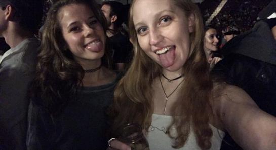 Two friends happily toast with drinks at a lively party surrounded by people.