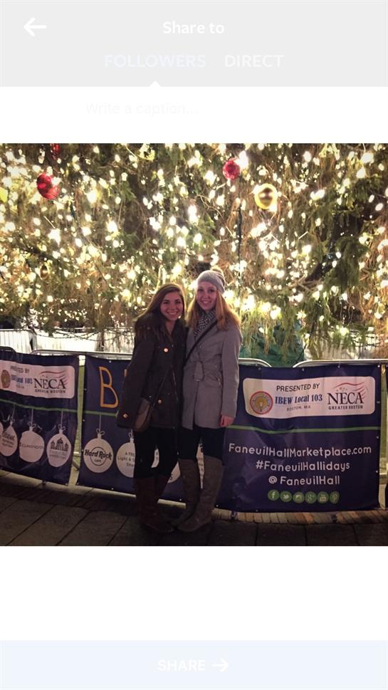 Two friends enjoy festive cheer at a holiday market near a brightly lit Christmas tree.