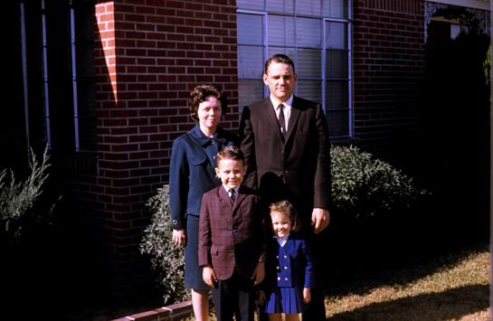 A family stands outside a brick building, smiling: a mother, father, and two young kids.
