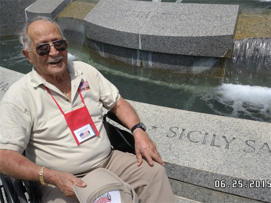 An elderly veteran smiles while sitting by a commemorative fountain dedicated to Sicily.