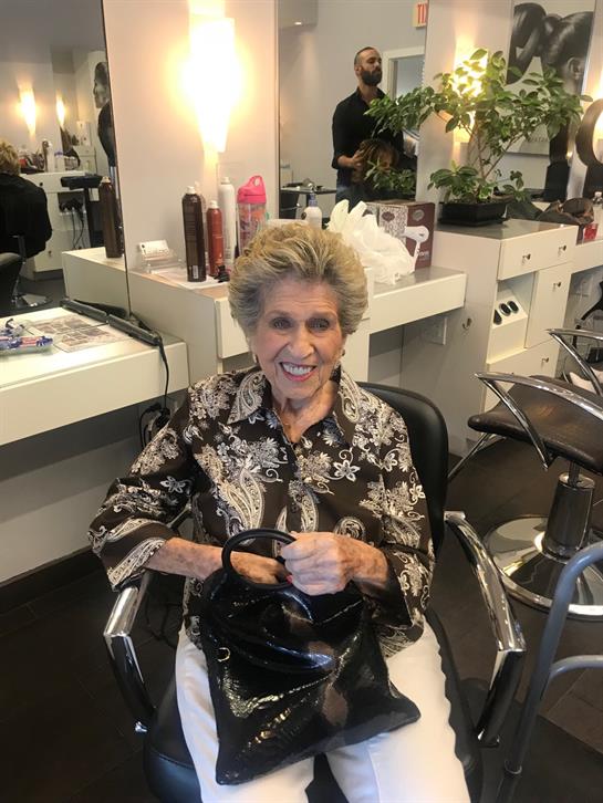 Elderly woman sits in a salon chair, smiling happily while waiting for her hair appointment.