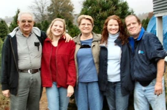 Five family members smile happily together outdoors at a tree farm surrounded by pine trees.