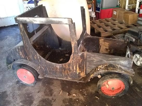 A handmade wooden toy car rests on a dusty floor in a workshop filled with tools and materials.