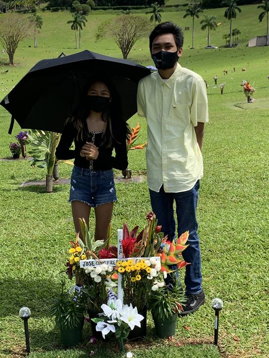Two friends stand together by a memorial site, holding an umbrella on a sunny day.