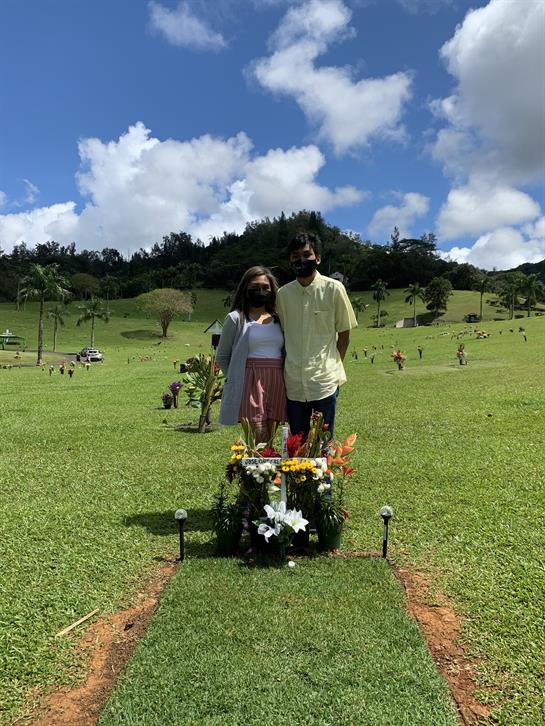 A couple remembers someone special at a sunny grave site surrounded by greenery.