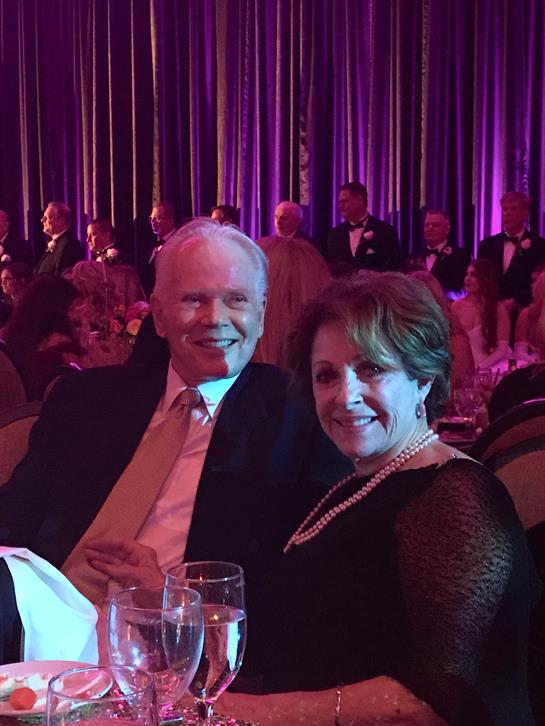 Couple poses happily at a formal gathering filled with guests and vibrant lighting.