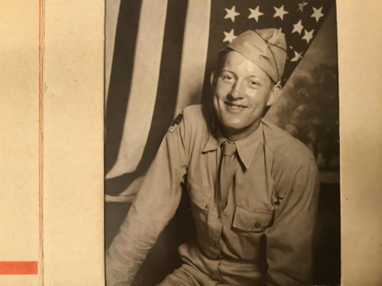 A smiling soldier sits in uniform, showcasing camaraderie and patriotism against a flag backdrop.