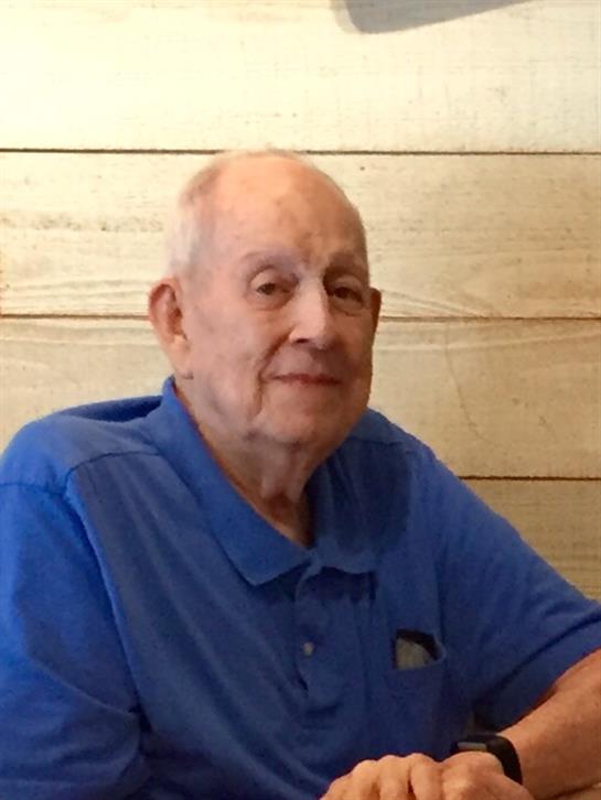 An elderly man sits casually at a wooden table, smiling and looking at the camera with warmth.