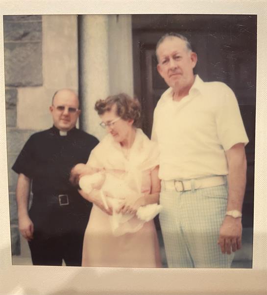 A priest and a couple hold an infant outside a historic building during the summer.
