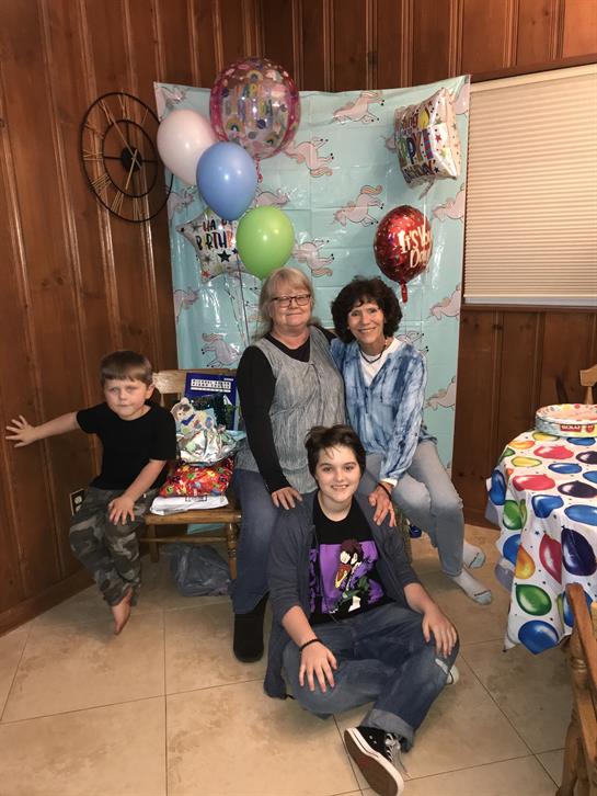 Family gathers in a warm, decorated space full of balloons and gifts, celebrating together joyfully.