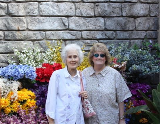 Two women smile among blooming flowers with a textured stone wall behind them.