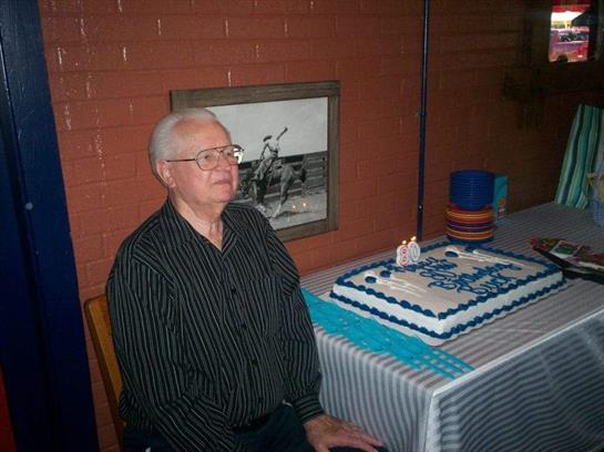 An elder man sits beside a beautifully decorated cake, surrounded by a festive yet calm atmosphere.