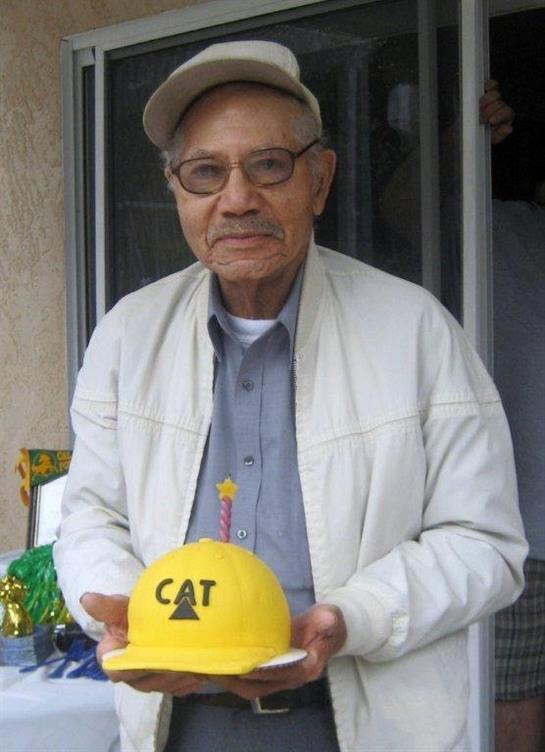 An elderly man in glasses and a cap smiles while holding a yellow cake with CAT lettering.