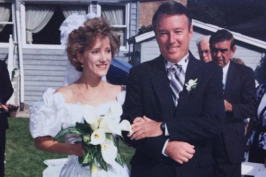 A smiling bride and groom stand together outside, surrounded by guests and flowers.