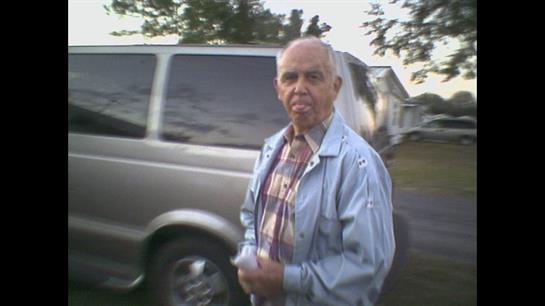 An elderly man, wearing a light blue jacket and plaid shirt, stands next to a silver van.
