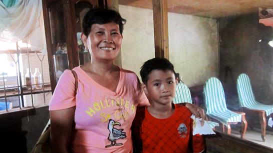A cheerful woman and a young boy stand side by side in a warm, inviting room with wooden accents.