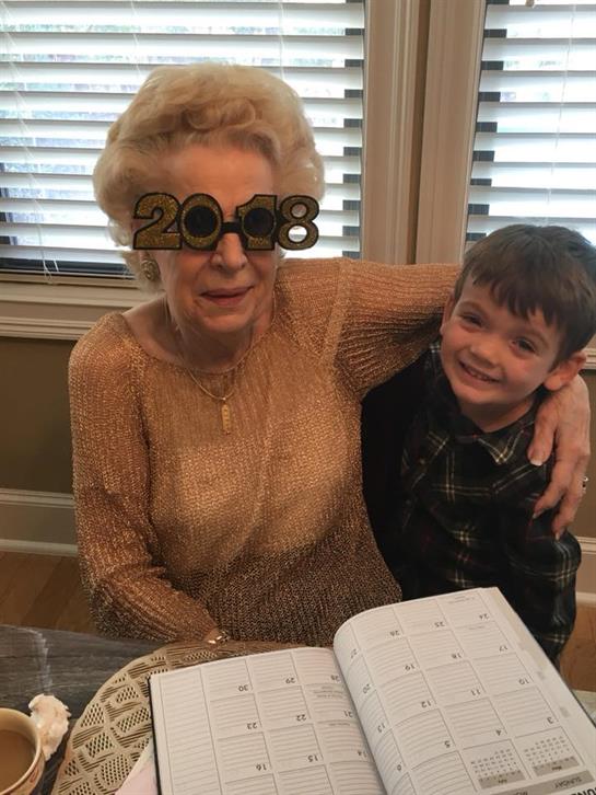 Grandmother wears novelty glasses while sitting beside her smiling grandson in a warm setting.