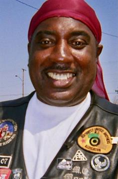 A joyful man dressed in a leather vest and bandana stands outdoors, exuding confidence and warmth.