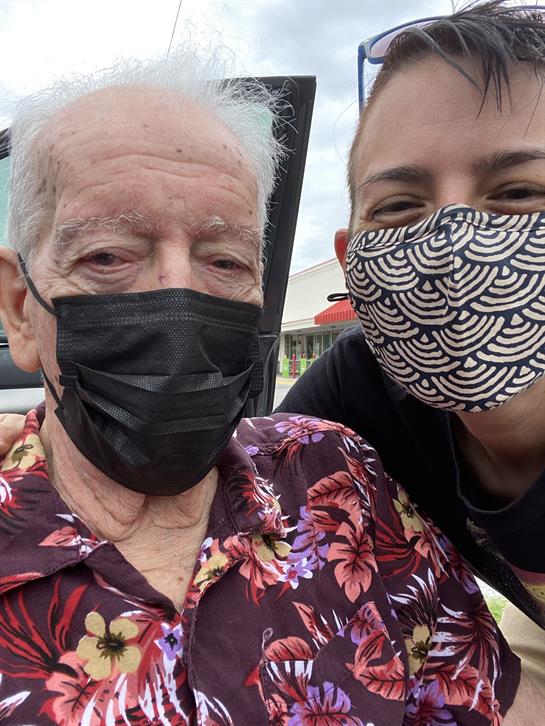 A senior man and a caregiver take a cheerful selfie outside, both wearing masks, enjoying the day.
