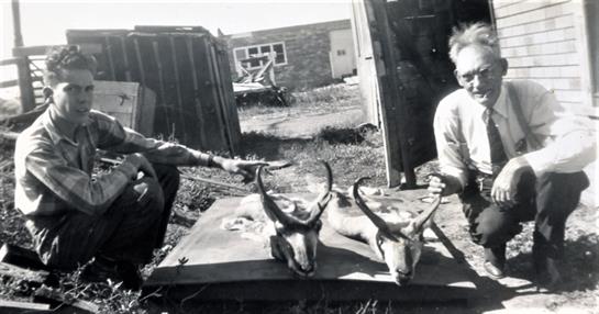 Two hunters pose with their catch of antelopes in a field behind a rustic building.