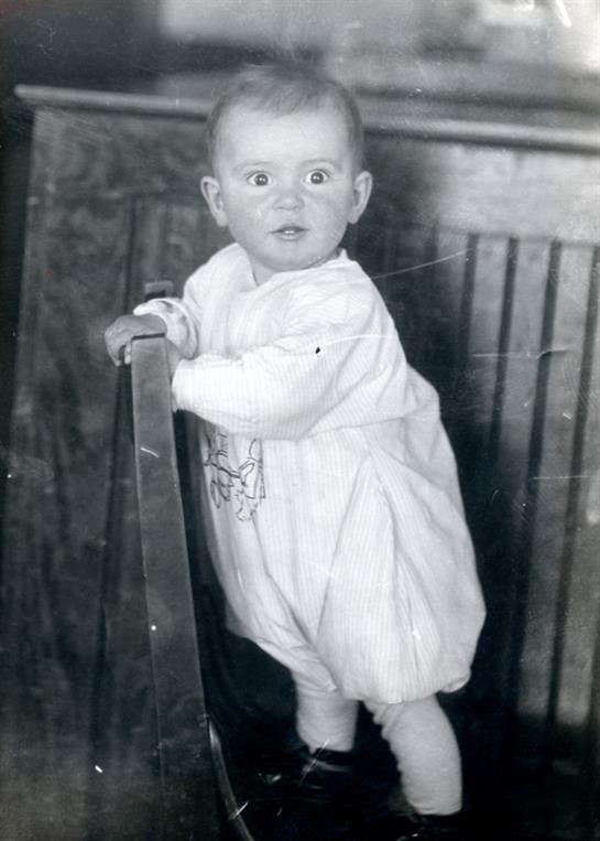 A curious child stands beside a wooden chair with wide eyes, dressed in a vintage outfit.