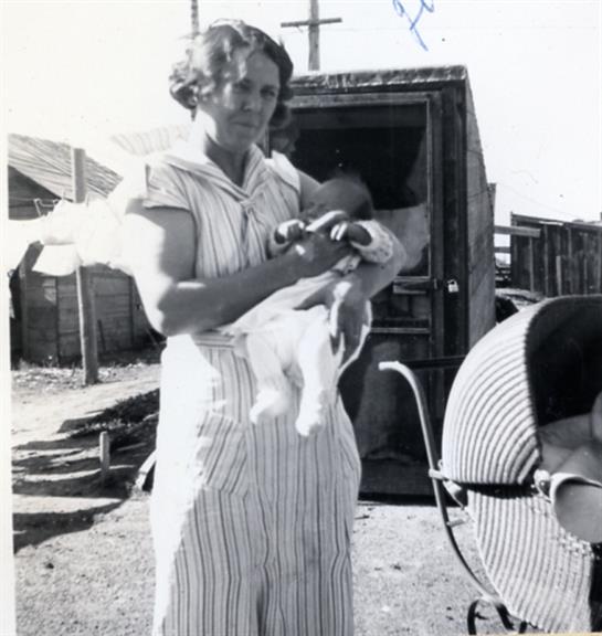 A woman dressed in light clothing holds a baby outside a modest home on a sunny day.
