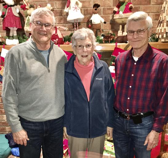 Three family members smile together in a warm living room decorated for the holiday season.