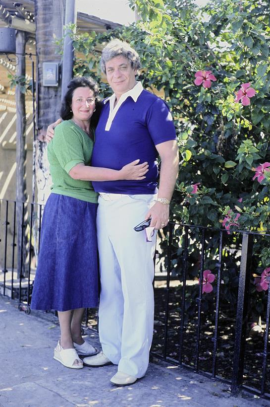 A couple stands closely together in a garden, enjoying the sunny weather and surrounding flowers.