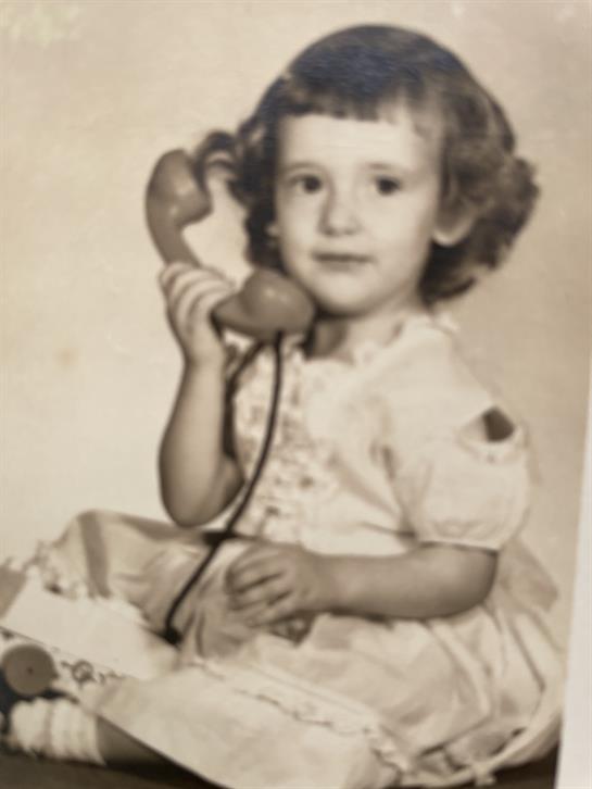 A young girl with curls smiles while holding a retro telephone, surrounded by childhood toys.