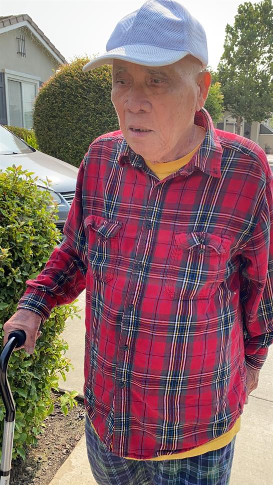 An elderly man uses a cane while walking along a pathway in a neighborhood filled with greenery.