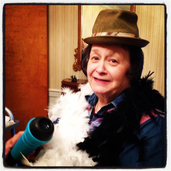 Woman dressed in a fun costume with a feather boa and hat smiles while holding a colorful prop.