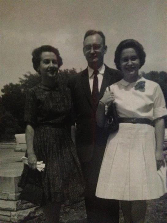 Three people stand close together, wearing formal outfits and smiling in a park setting.