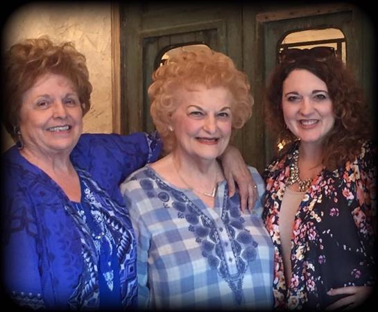 Three women smile brightly, embracing each other in a warm indoor space filled with joyful energy.
