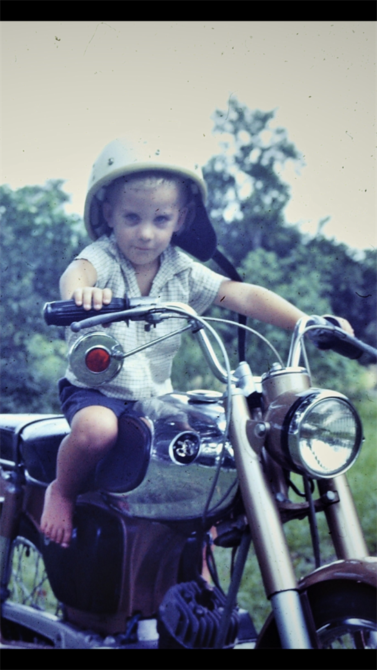 A child in a helmet confidently rides a classic motorcycle amidst green trees and bright sunshine.