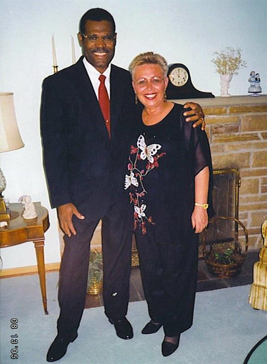Two friends stand side by side, smiling in a warmly lit living room with a fireplace.