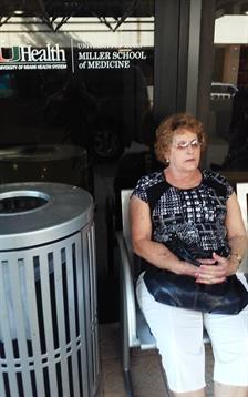 A senior woman in stylish glasses sits calmly beside a trash can in a busy area during daylight.