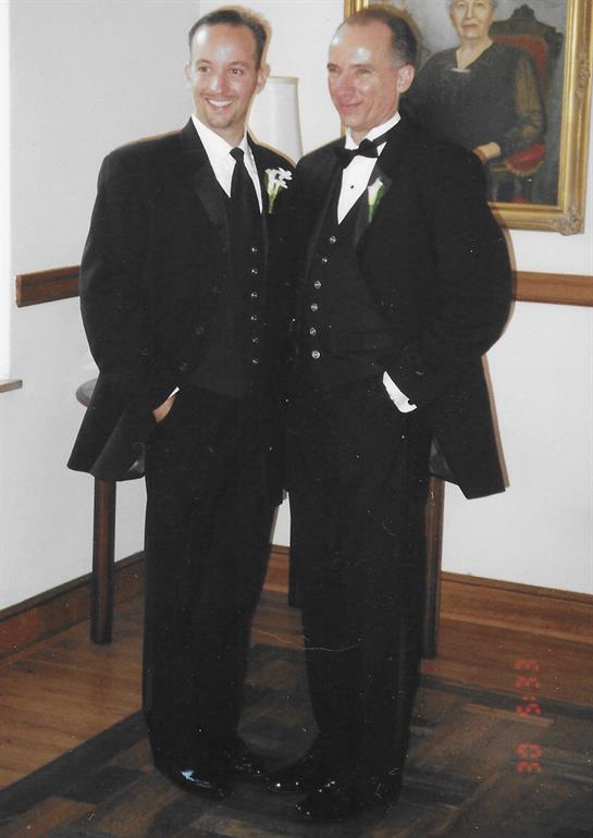 Two well-dressed men stand side by side, dressed in black suits, in a well-lit room.